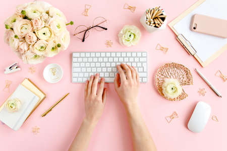 Stylized, pink womens home office desk. Workspace with female hands, computer, bouquet ranunculus...