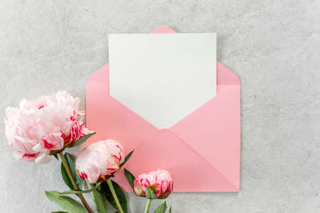 Mockup invitation, blank paper greeting card, pink envelope and peonies on gray stone table. Flower...