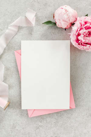 Mockup invitation, blank paper greeting card, pink envelope and peonies on gray stone table. Flower background. Flat lay, top view.の写真素材
