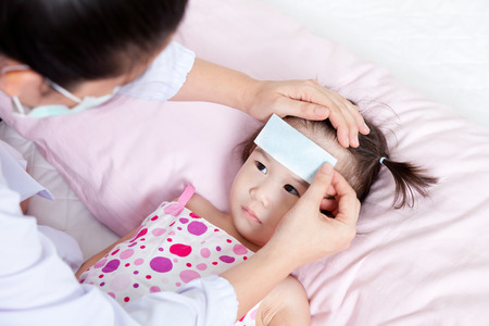 Portrait of confident female doctor paste cooling fever patch on forehead little asian girl during medical treatment, girl lying on sickbedの写真素材