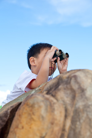 Happy little asian boy exploring outdoors prone on a boulder with telescope, young boy using binoculars to view the front, bright sky backgroundの写真素材