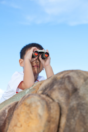 Happy little asian boy exploring outdoors prone on a boulder with telescope, young boy using binoculars to view the front, bright sky backgroundの写真素材