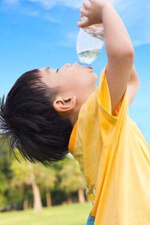 Little asian boy drinking water from plastic bottle with the thirsty, after tired from a romp in the park, under bright sunlight and blue sky background, outdoor shotの写真素材