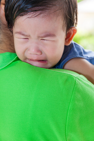 Mother carrying and comfort her daughter in backyard over blur green nature background, little asian girl (thai) crying on her mother shoulderの写真素材