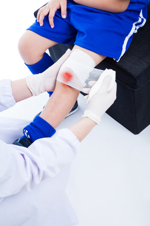 Youth asian (thai) sport boy in blue uniform. Doctor perform first aid knee injure by bandage, bloody on bandage. Shoot in studio. On white backgroundの写真素材