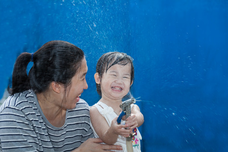 Funny laughing little asian (thai) girl and her mother playing with water hose at the day time, on blue background, loving and happy familyの写真素材