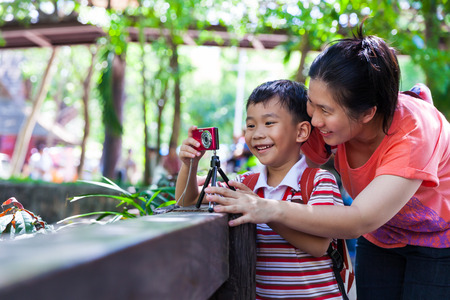 Portrait of young woman teaching little asian (thai) boy photographing outdoors using digital camera. Happy family, Mother and son smiling together in the parkの写真素材