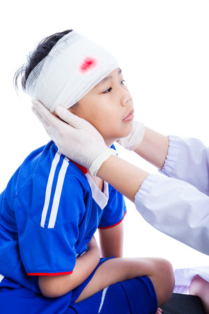 Sports injury. Athlete little asian (thai) boy in blue sportswear with trauma of the head, bloody on bandage. Doctor makes a bandage on head patient, on white background. Studio shoot. Side view.の写真素材