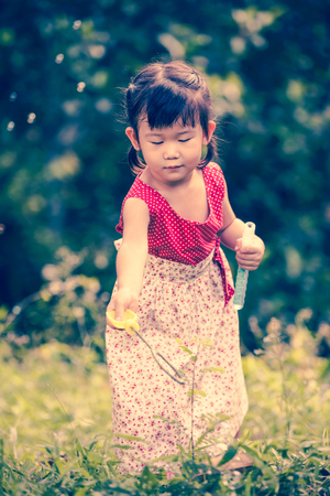 Asian lovely girl with beautiful dress playing on grass in the park. Happy child reaching for soap bubbles on blurred nature background. Outdoors. Vintage picture style.の写真素材