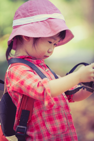 Cute little asian girl checking and reviewing photos in professional digital camera over nature background. Active lifestyle, curiosity, pursuing a hobby, technology and kids concept. Retro style. Outdoors.の写真素材