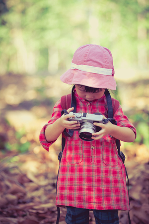 Cute little asian girl checking and reviewing photos in professional digital camera over nature background. Active lifestyle, curiosity, pursuing a hobby, technology and kids concept. Retro style. Outdoors.の写真素材