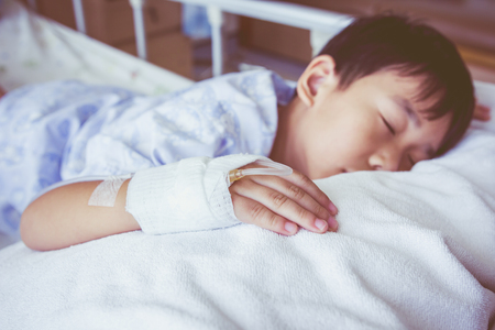 Illness asian boy sleeping on sickbed in hospital, saline intravenous (IV) on hand, shallow depth of field (DOF) saline intravenous (IV) in focus, child out of focus. Retro style.の写真素材