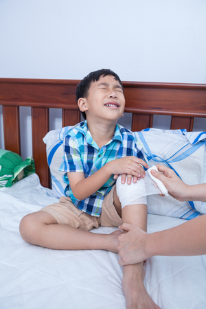Child injured. Mother bandaging son's knee on bed inside bedroom, bandage in focus, asian boy sadden and crying. Human health care and medicine concept.の写真素材
