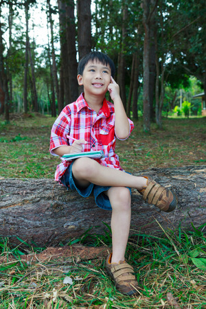 Handsome asian boy thinking to solve a problem on wooden log in national park. Outdoors in the day time with bright sunlight. Children planning and education concept.の写真素材