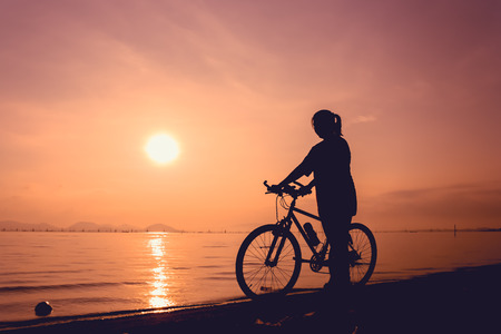 Silhouette of healthy biker-girl enjoying the view at seaside, on colorful sunset orange sky background. Reflection of sun in water. Active outdoor lifestyle concept. Outdoors.の写真素材