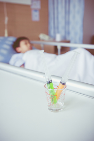 Syringes in a glass measuring cup with blurred illness boy lying on sickbed in hospital. Syringe in focus, child out of focus. Health care and medical concept. Vintage style.の写真素材