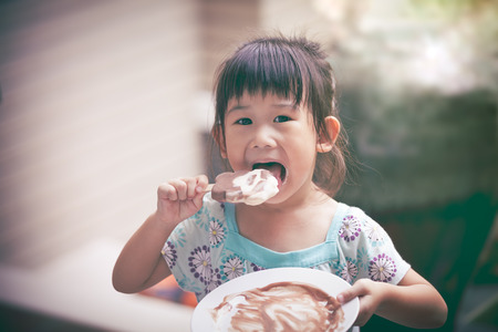 Pretty asian girl eating ice cream in the summer on blurred background. Happy child looking at camera. Outdoors. Vintage style.の写真素材