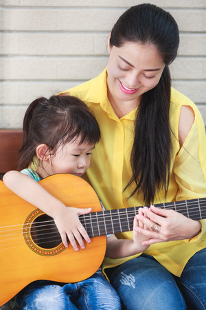 Happy family spending time together at home. Asian mother with daughter playing classic guitar. Positive human emotion.の写真素材