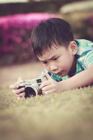 Asian boy taking photo by vintage film camera on blurred nature background at the day time. Adorable child enjoying at park. Outdoors. Vintage picture style.の写真素材
