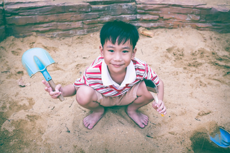 Happy child playing with sand and smiling. Adorable asian boy has fun digging in the sand on a summer day and looking at camera. Vintage tone. Outdoors.の写真素材