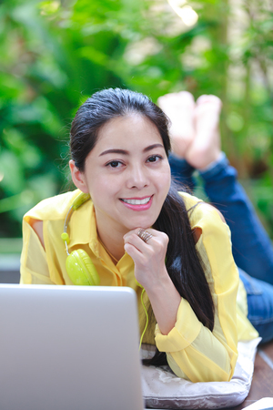 Smiling asian woman using laptop computer while lying on floor with earphones for entertainment. Female working on laptop and looking at camera. Outdoors.の写真素材