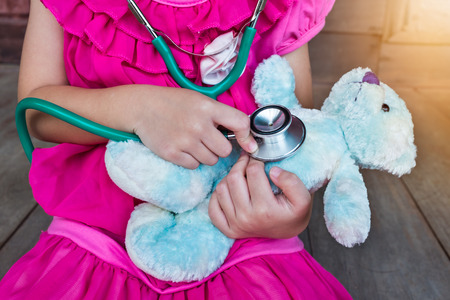Child playing doctor or nurse with plush toy bear with bright sunlight at home. Happy girl listens a stethoscope to toy. Playful girl role playing.の写真素材