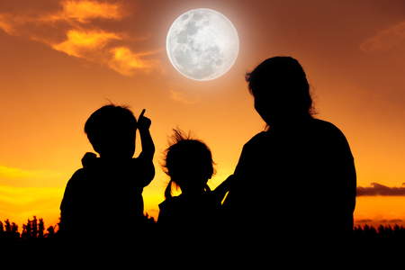 Silhouette back view of family sitting and relaxing together. Boy point to full moon on gold sky background. Happy family spending time together. The moon taken with my own camera, no NASA images usedの写真素材