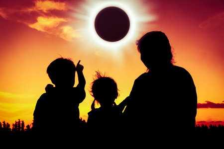 Silhouette back view of family sitting and relaxing together. Boy point to solar eclipse on gold sky background. Happy family spending time together. Outdoor.の写真素材