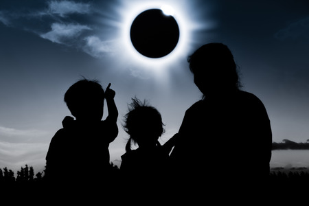 Natural phenomenon. Silhouette back view of mother and child sitting and relaxing together. Boy pointing to solar eclipse on dark sky background. Happy family spending time together. Outdoor.の写真素材
