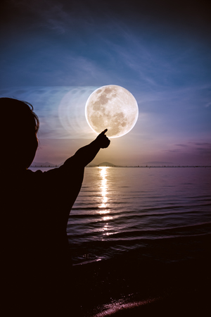 Silhouette back view of woman relax at seaside and pointing to the full moon on colorful sky background. Outdoors. Abstract full moon movement. The moon were NOT furnished by NASA.の写真素材