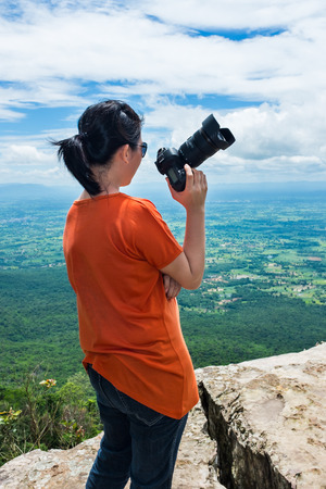 Asian woman with digital camera at view point on the top of mountain, travel on vacation. Back view of woman standing against sky with cloudy. Outdoor at the daytime on summer day.の写真素材