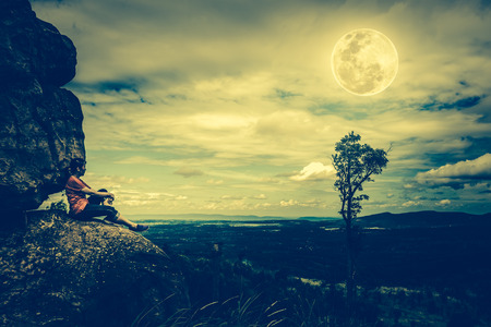Woman sitting on boulders and looking at  sky with cloudy and beautiful full moon over tranquil nature nighttime. Low key and cross process tone effect.の写真素材