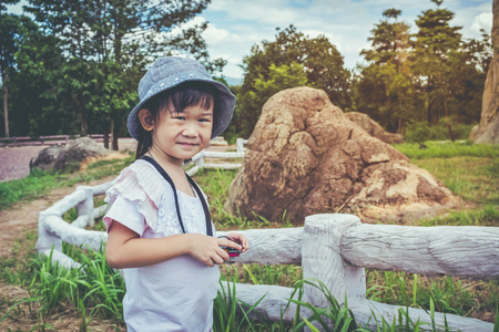 Happy child photographer with hat smiling and holding camera at park. Asian girl shooting pictures of nature. Pretty child relaxing outdoors on summer day, travel on vacation. Vintage effect tone.の写真素材