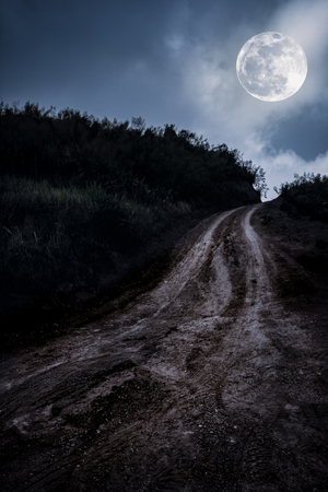 Landscape in nature of beautiful full moon with tire tracks on a muddy road through a forest. Mountain winding road passing, outdoors at nighttime. Dark tone.の写真素材