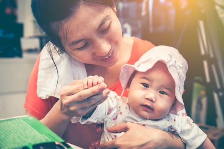 Lovely asian baby girl sitting on mother lap. Happily mom smiling and take care to comfort her daughter with love at home. Happy family spending time together. Vintage tone. Mother's Day celebration.の写真素材
