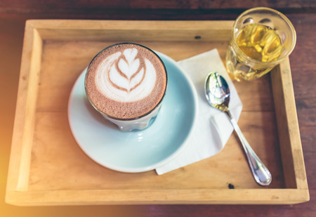 Top view cup of hot cocoa art heart-shape and leaf figure milk foam on saucer with spoon and herbal tea. Beverages placed in a tray on a wooden table with bright sunlight.の写真素材