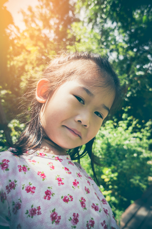 Adorable asian child smiling and relaxing at park on blurred nature background. Happy girl looking at camera. Outdoors with bright sunlight on summer day, travel on vacation. Vintage tone.の写真素材