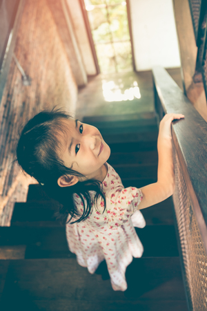 Adorable asian girl in beautiful dress coming down wooden stairs and looking back at home. Happy child looking at camera. Daytime with bright sunlight. Vintage effect tone.の写真素材