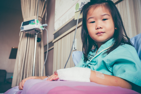 Illness asian child looking at camera, admitted in hospital with infusion pump and saline intravenous (IV) on hand. Girl feeling sad. Health care stories. Vintage film filter effect.の写真素材