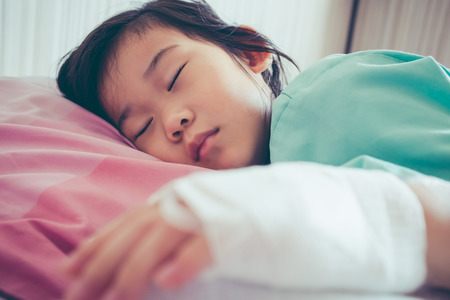 Closeup of illness asian child admitted in hospital with saline intravenous (IV) on hand. Girl sleeping at comfortable equipped hospital room. Health care stories. Vintage film filter effect.の写真素材