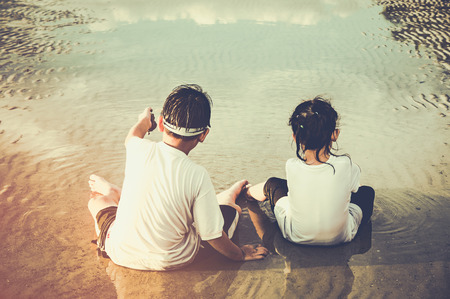 Back view of asian brother and sister playing with sand. Happiness siblings enjoy on the beach. Outdoors with sunlight on summer day. Concept of connecting children with nature. Travel on vacation.の写真素材