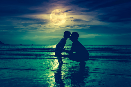 Father's day. Silhouette of loving child kissing her father. Family enjoying and relaxing on beach with full moon on sky background in the evening. Cross process. The moon taken with my own camera.の写真素材