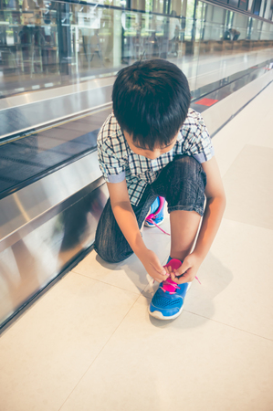 Asian child sitting and tying to tie shoelaces near electric speedwalk in modern airport. Do it yourself concept. Travel on vacation. Vintage film filter effect.の写真素材