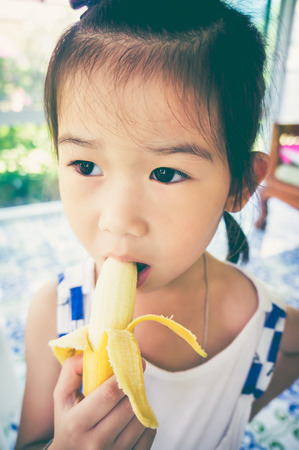 Close up adorable asian girl peeling and eating a ripe banana at  home.  Child healthy food eating concept. Vintage film filter effect.の写真素材