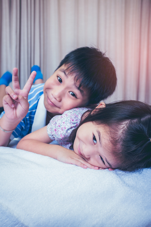 Lovely asian children relaxing in bedroom at home. Brother showing v-sign and sister smiling with sunlight. Happy family spending time together, loving and bonding of sibling. Vintage film effect.の写真素材