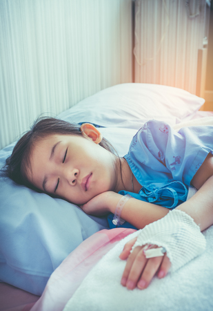 Illness Asian child admitted in hospital with saline intravenous (IV) on hand. Girl sleeping at comfortable equipped hospital room with sunlight. Health care stories. Vintage film filter effect.の写真素材