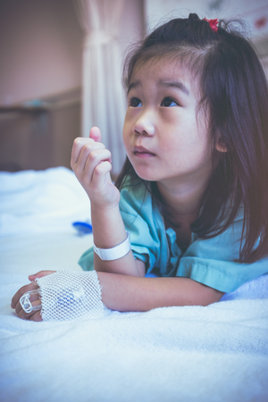 Illness asian child admitted in hospital, saline intravenous (IV) on hand. Adorable girl lie in prone on sickbed. Health care stories. Vintage film filter effect.の写真素材