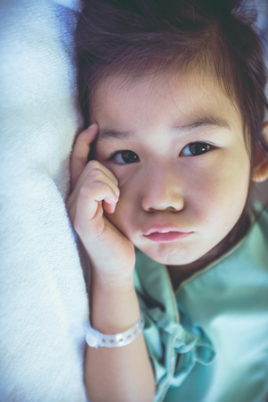 Closeup illness asian child lying on sickbed and looking at camera, admitted in hospital. Unhappy girl feeling bored and sad. Health care stories. Vintage film filter effect.の写真素材