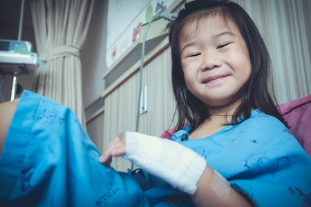 Illness asian child smiling happily and looking at camera. Young pretty girl admitted in hospital while saline intravenous (IV) on hand. Health care stories. Vintage film filter effect.の写真素材
