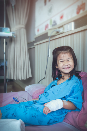 Illness asian child smiling and looking at camera. Young pretty girl admitted in hospital while saline intravenous (IV drip) on hand. Health care stories. Vintage film filter effect.の写真素材